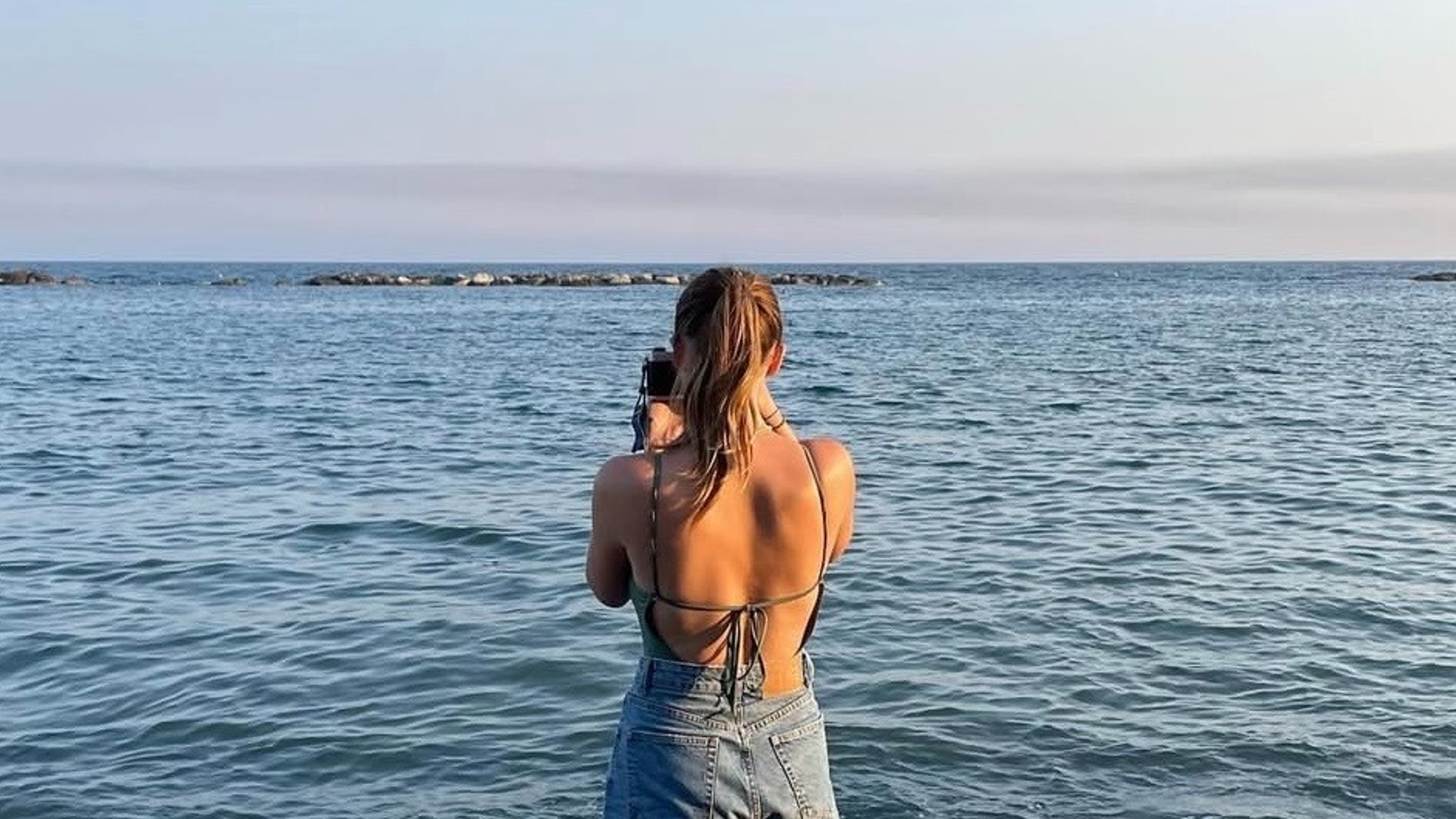 A girl stood on the beach looking out to sea holding a film camera and taking a picture.