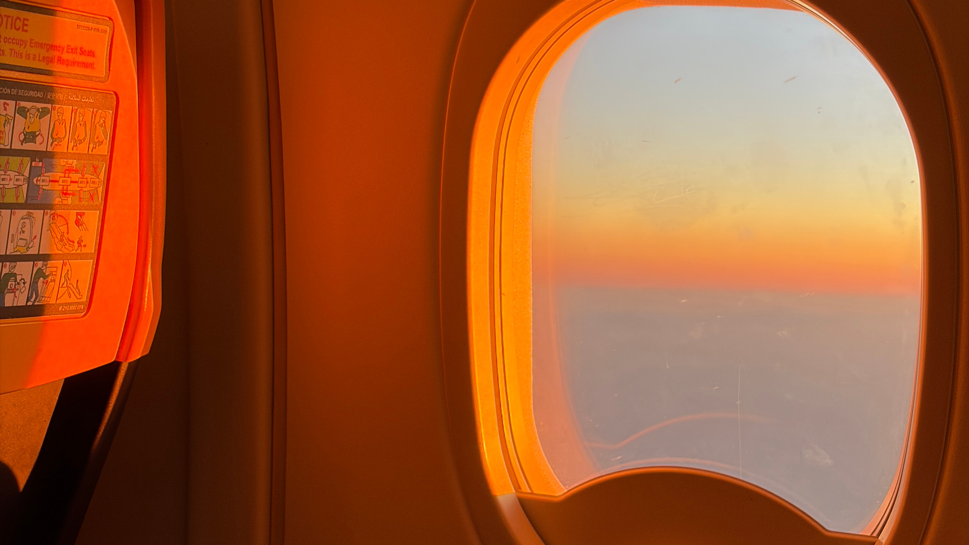 Photo of a plane window at golden hour looking out at the clouds.