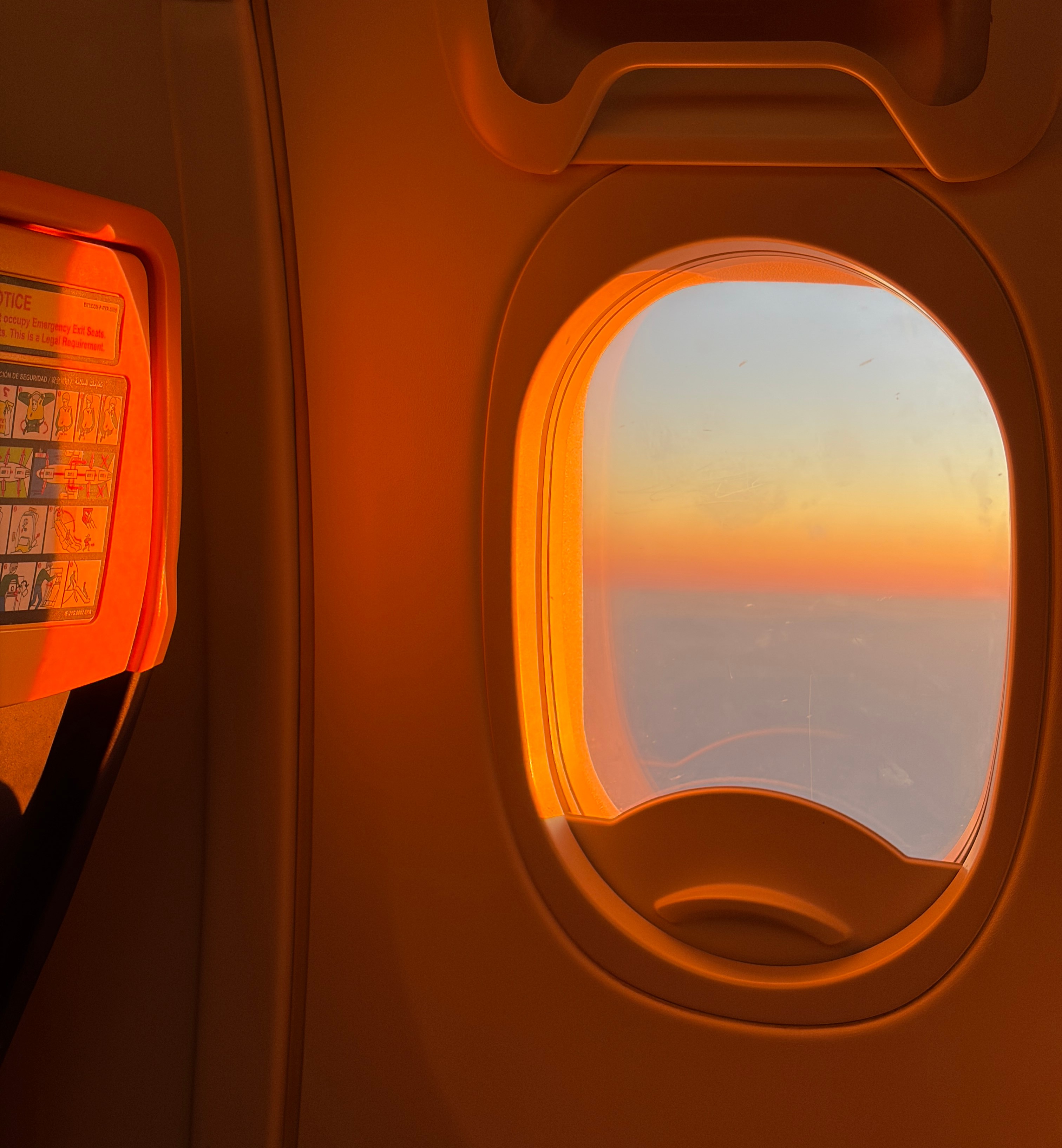 Photo of a plane window at golden hour looking out at the clouds.
