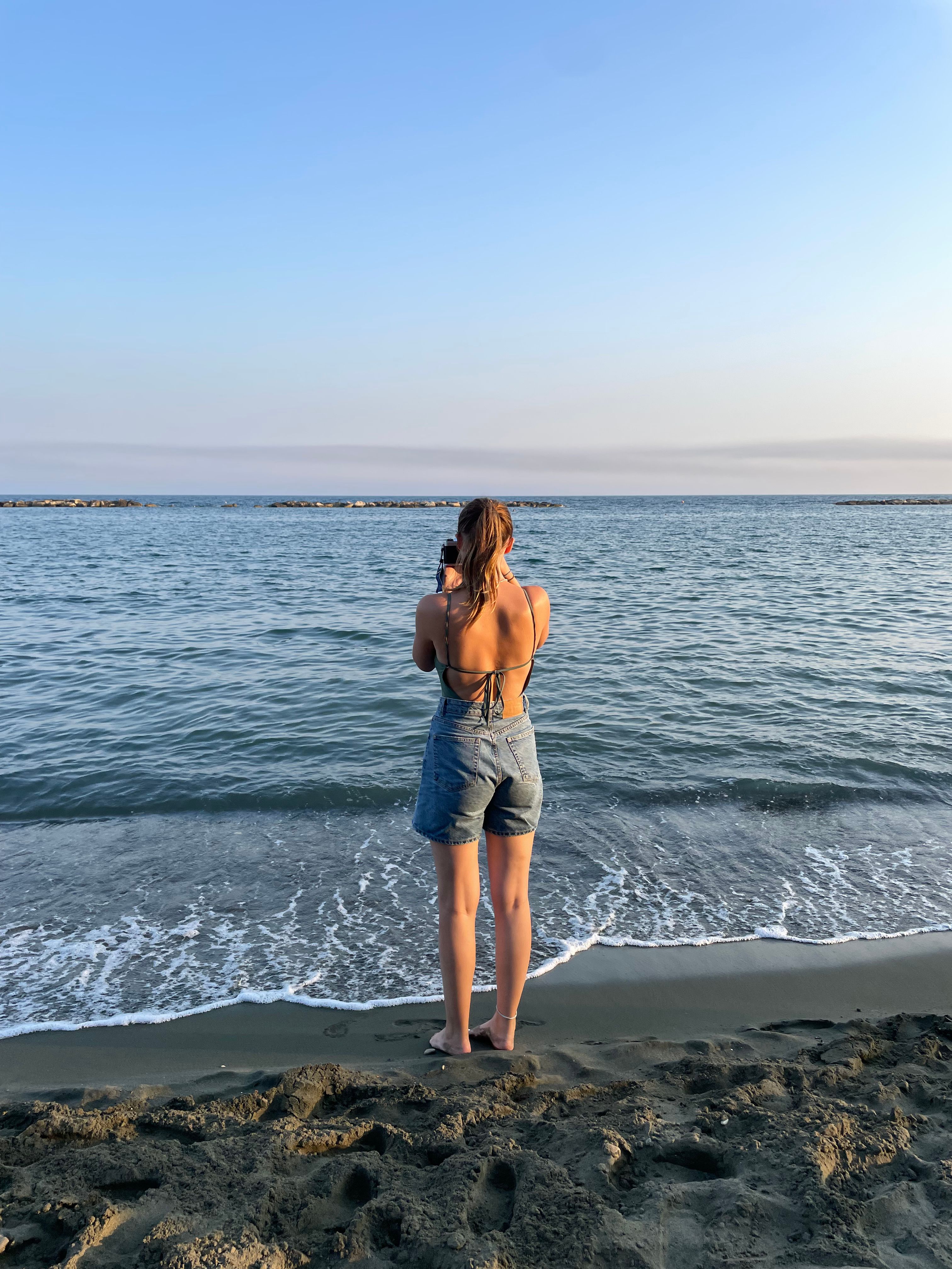 A girl stood on the beach looking out to sea holding a film camera and taking a picture.
