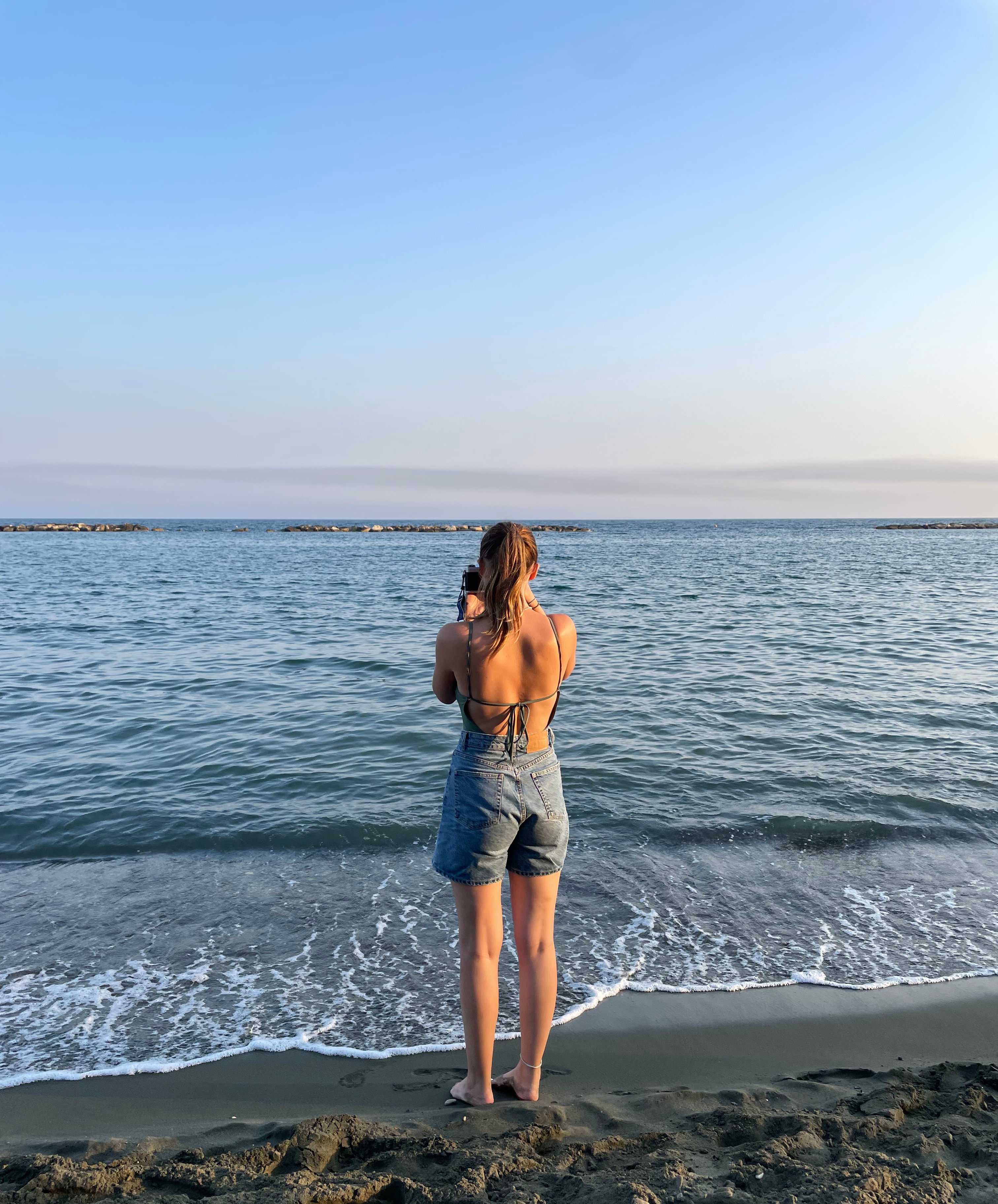 A girl stood on the beach looking out to sea holding a film camera and taking a picture.