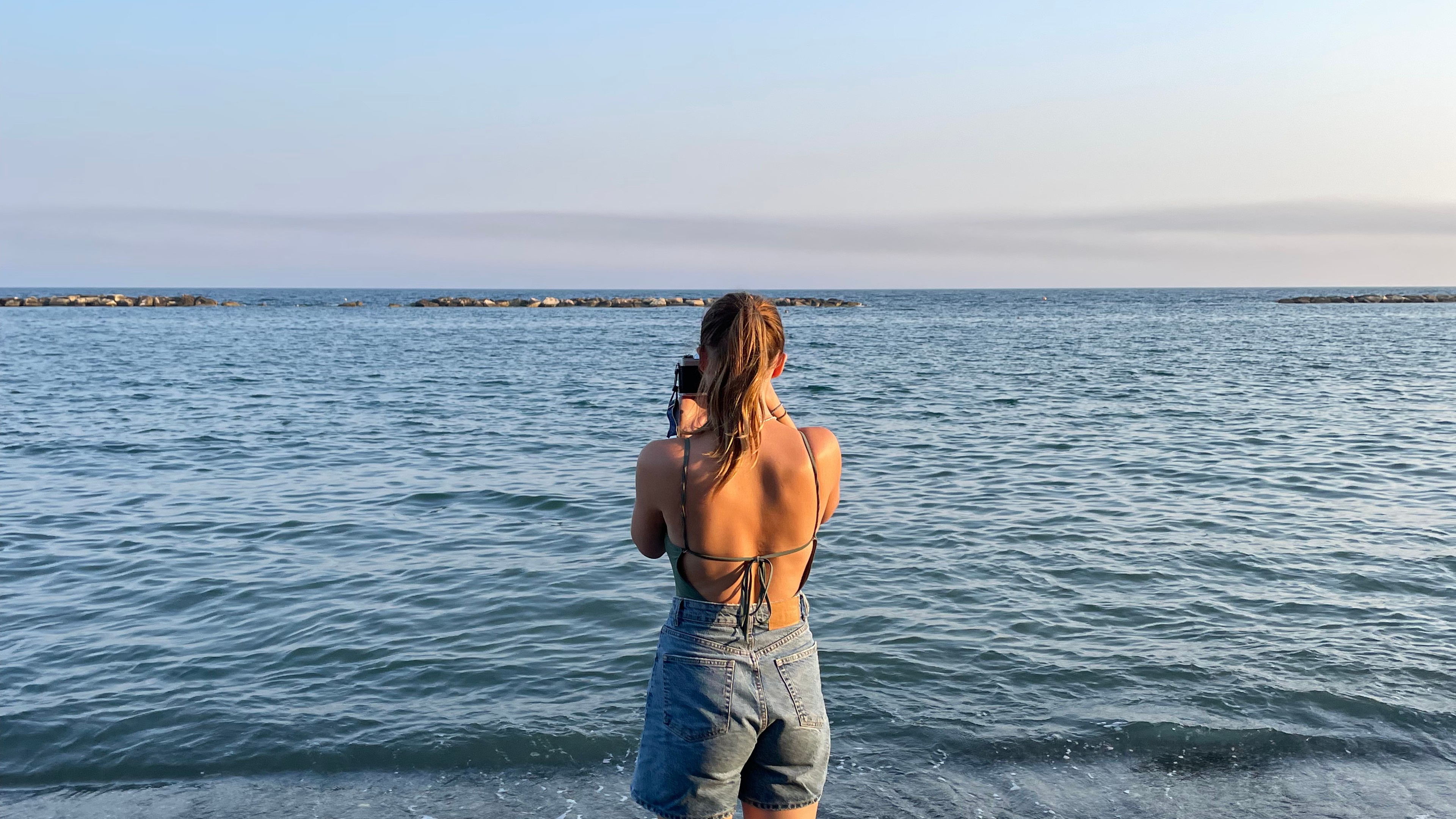 A girl stood on the beach looking out to sea holding a film camera and taking a picture.