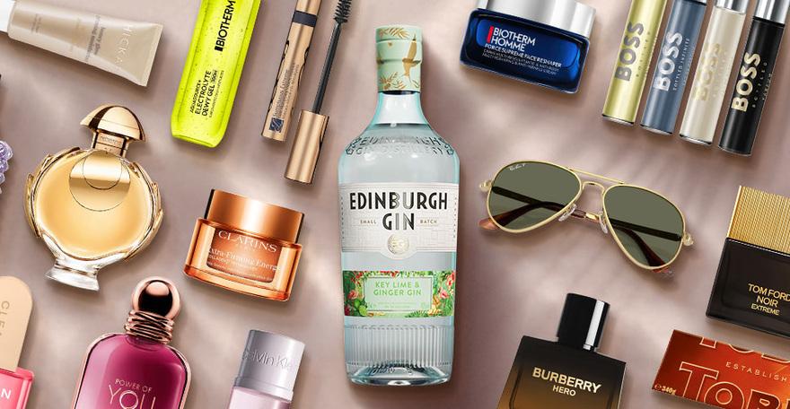 A flat lay of beauty products, perfumes, a bottle of gin, sunglasses, and a chocolate bar.