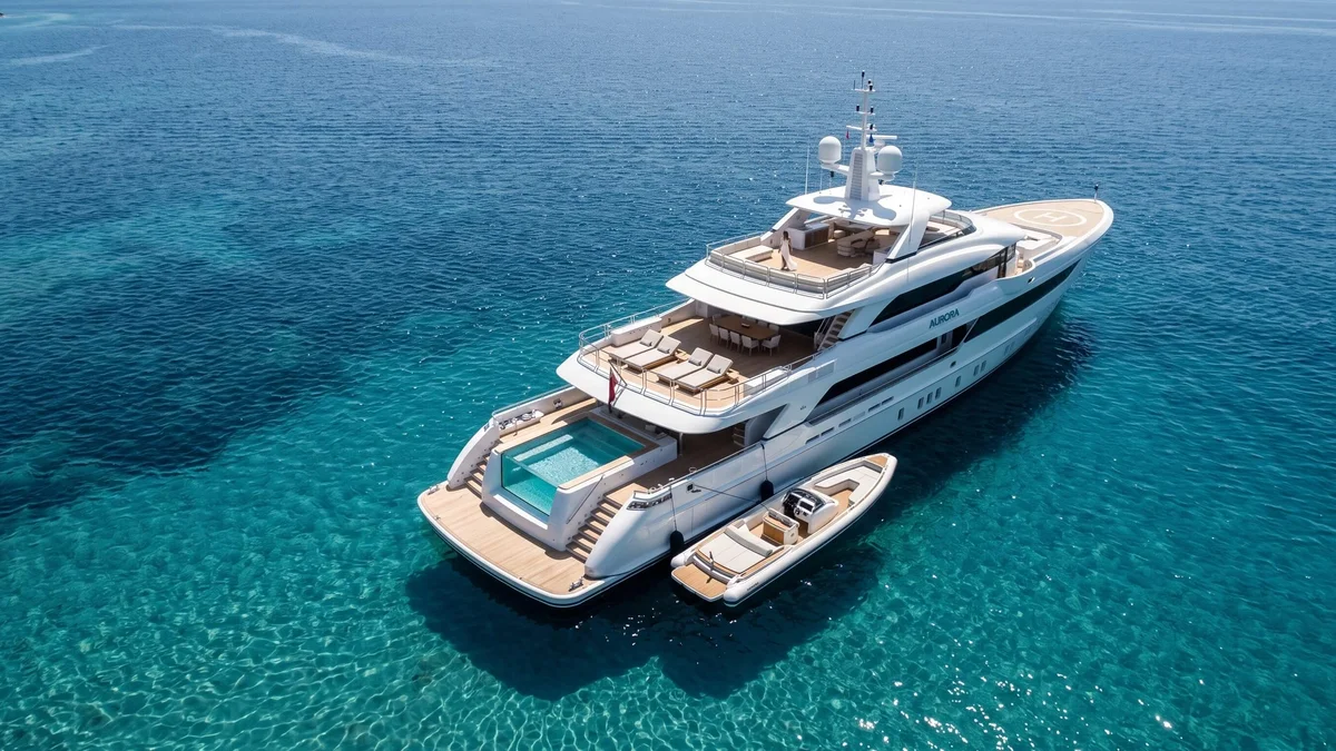 High-angle aerial view of a luxury white superyacht with a glass-walled pool and tender boat anchored in clear turquoise water.