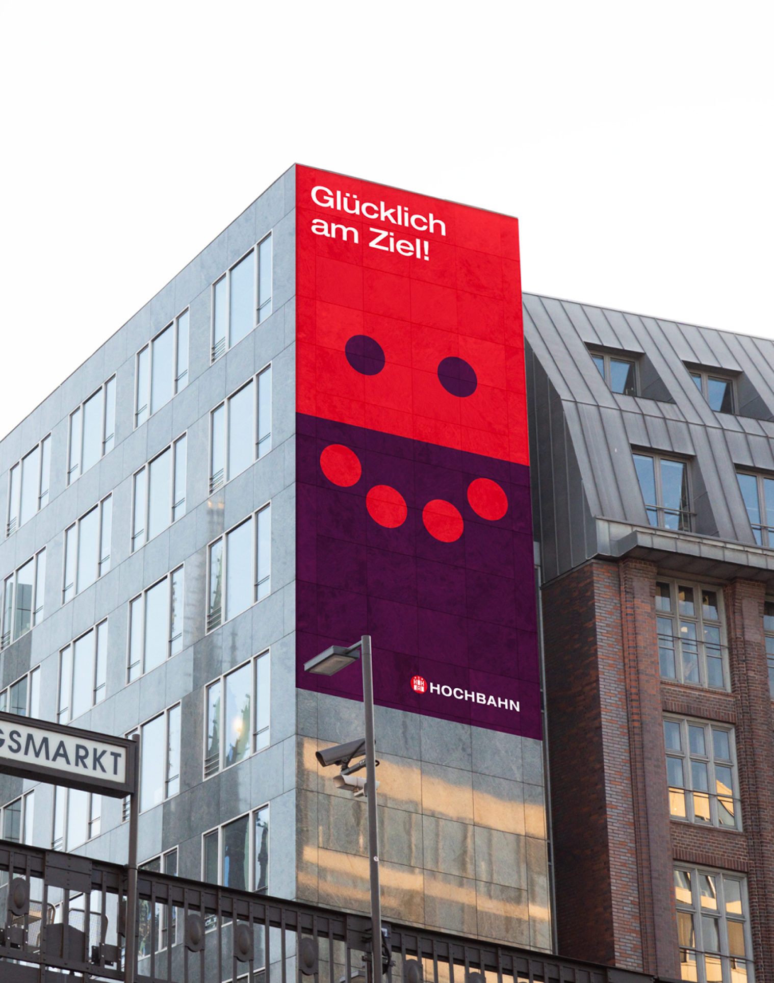 Hochbahn-Plakat an einer Hausfassade: Rote und violette Fläche bilden ein lächelndes Gesicht mit Claim „Glücklich am Ziel!".