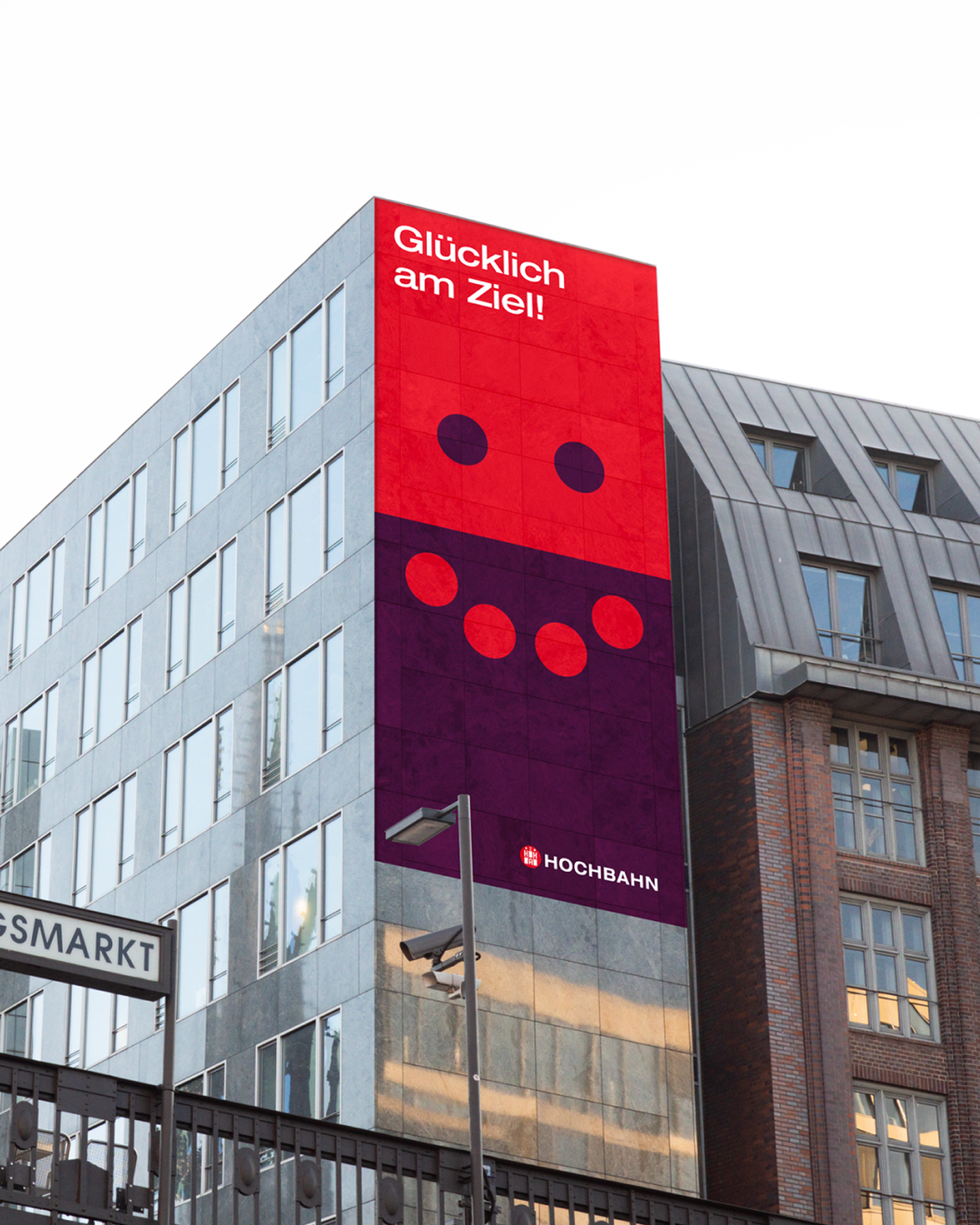 Hochbahn-Plakat an einer Hausfassade: Rote und violette Fläche bilden ein lächelndes Gesicht mit Claim „Glücklich am Ziel!".
