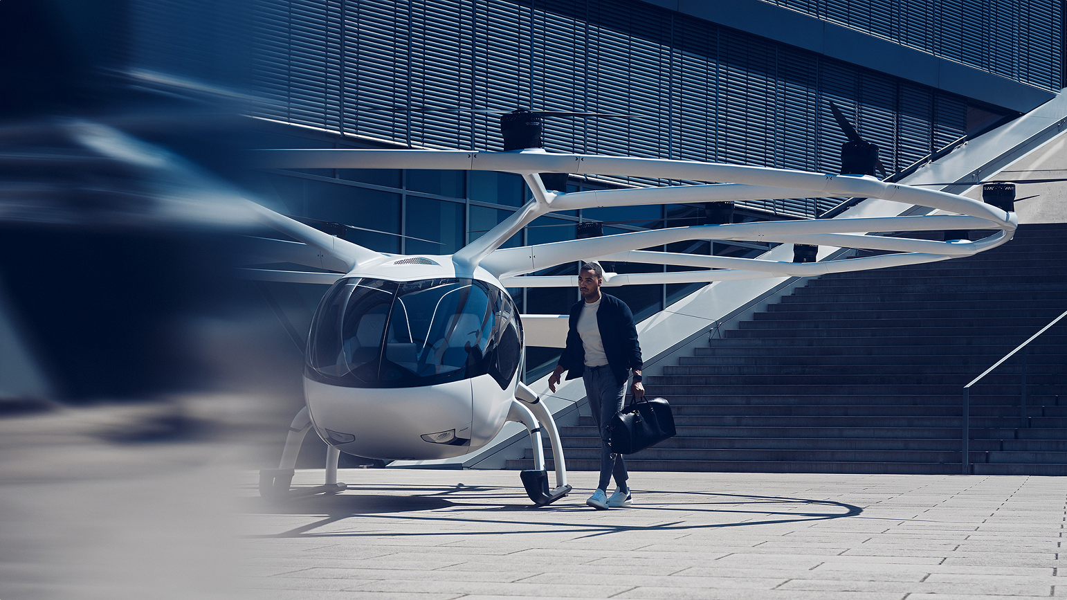 Volocopter auf urbanem Platz im Sonnenlicht; Mann mit Aktentasche läuft vorbei; Rotor in Bewegungsunschärfe