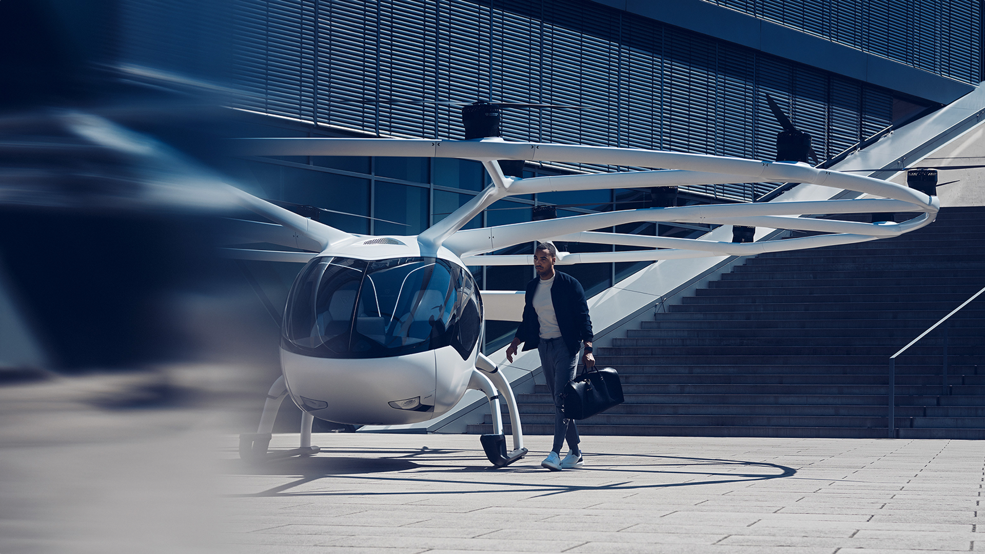 Volocopter auf urbanem Platz im Sonnenlicht; Mann mit Aktentasche läuft vorbei; Rotor in Bewegungsunschärfe