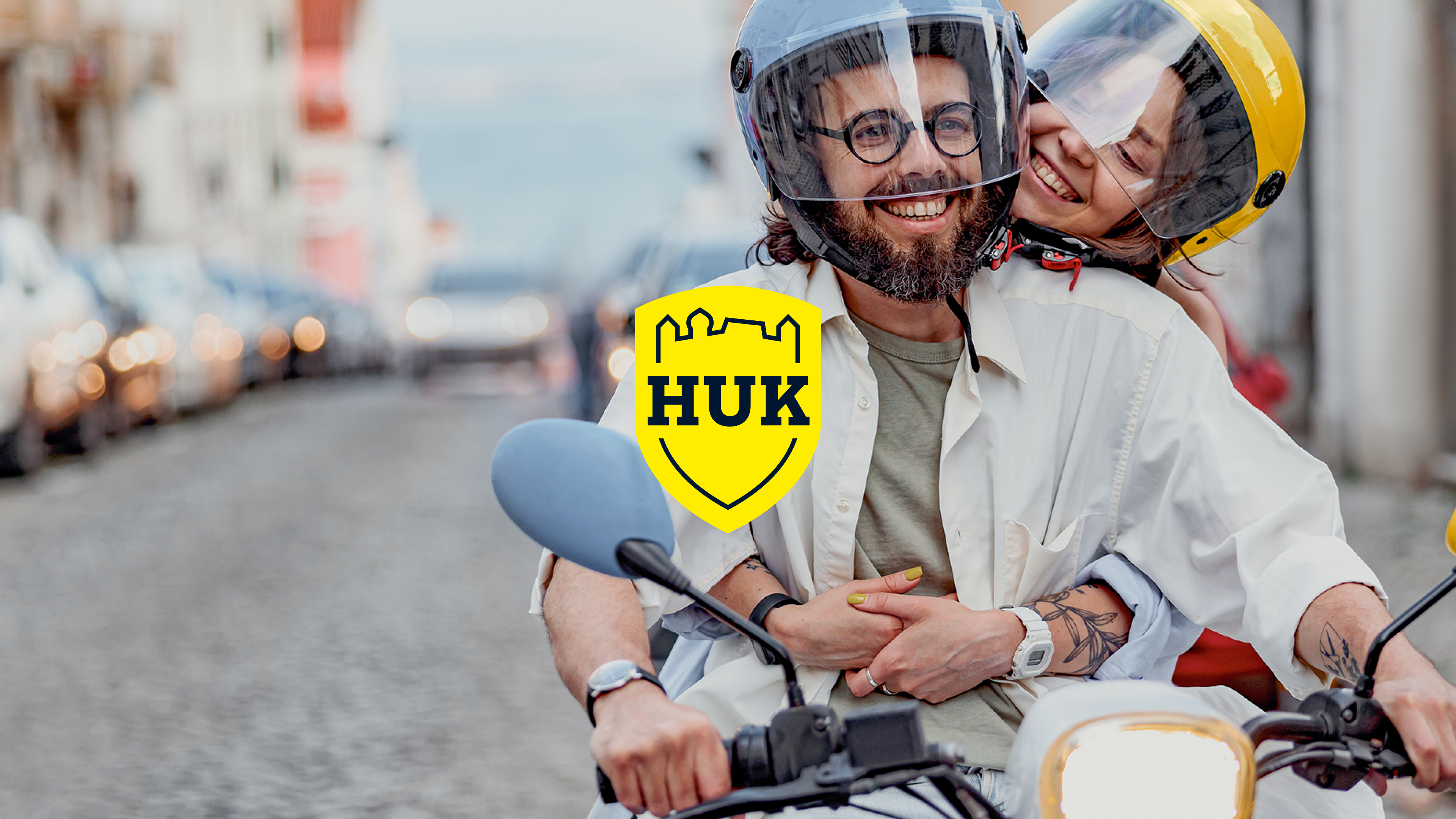 Ein lächelndes Paar mit Helmen auf einem Motorroller in einer Stadtstraße; im Vordergrund das gelbe HUK-COBURG Logo als Overlay.