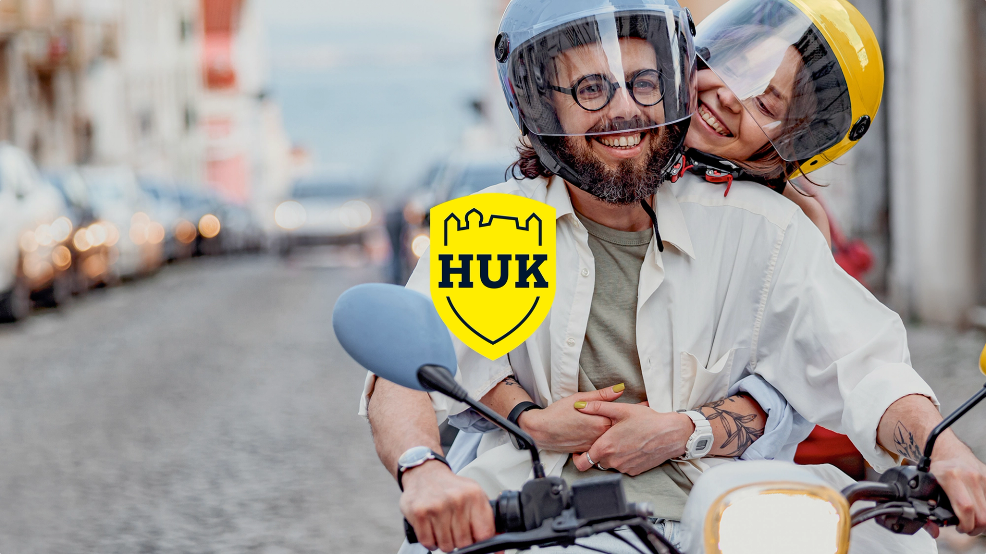 Ein lächelndes Paar mit Helmen auf einem Motorroller in einer Stadtstraße; im Vordergrund das gelbe HUK-COBURG Logo als Overlay.