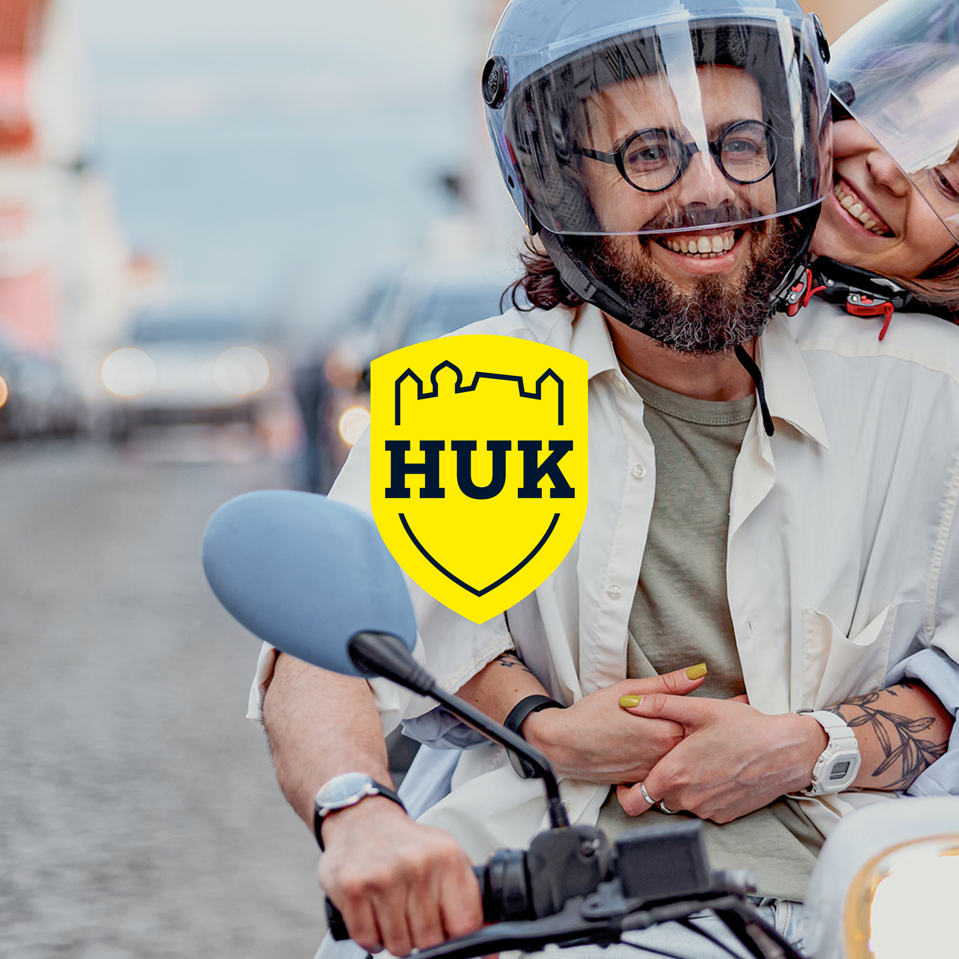 A smiling couple wearing helmets on a scooter on a city street; in the foreground, the yellow HUK-COBURG logo as an overlay.