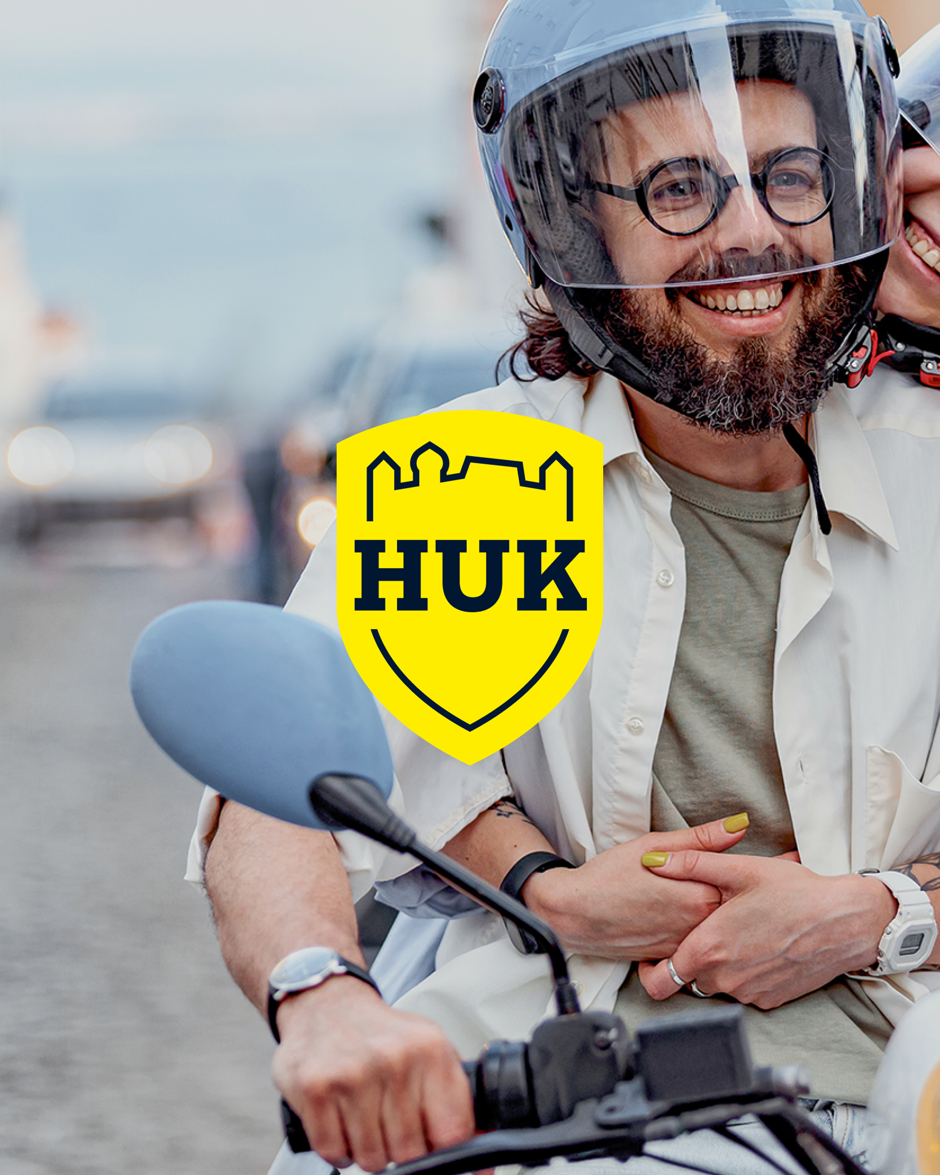 Ein lächelndes Paar mit Helmen auf einem Motorroller in einer Stadtstraße; im Vordergrund das gelbe HUK-COBURG Logo als Overlay.