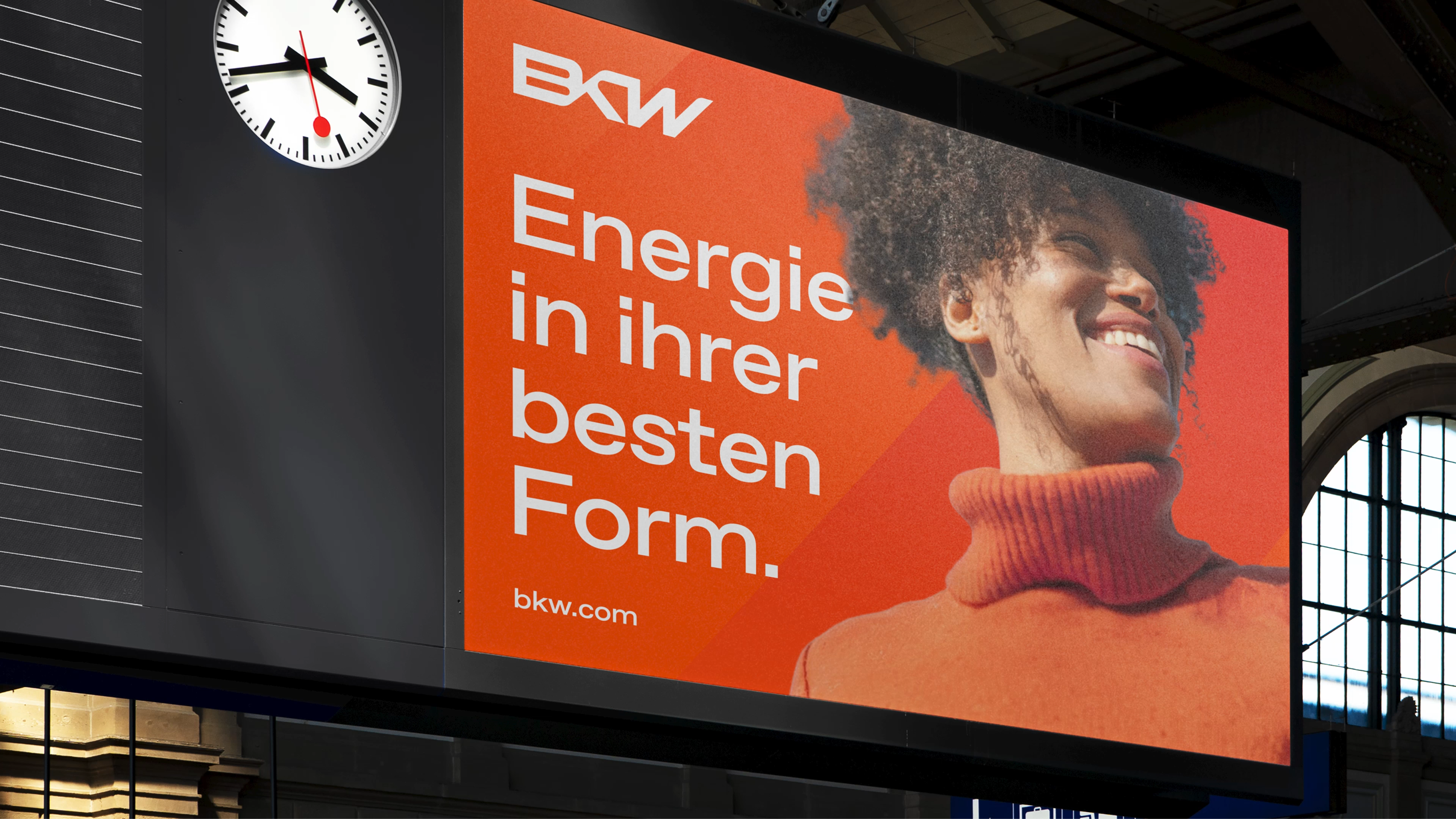 BWK-Plakat im Bahnhof: lächelnde Person vor orangefarbenem Grund, Text „Energie in ihrer besten Form.“ bkw.com