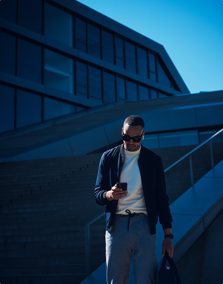 Mann mit Sonnenbrille und Tasche blickt auf Smartphone, blau getöntes Gegenlicht, urbane Architektur