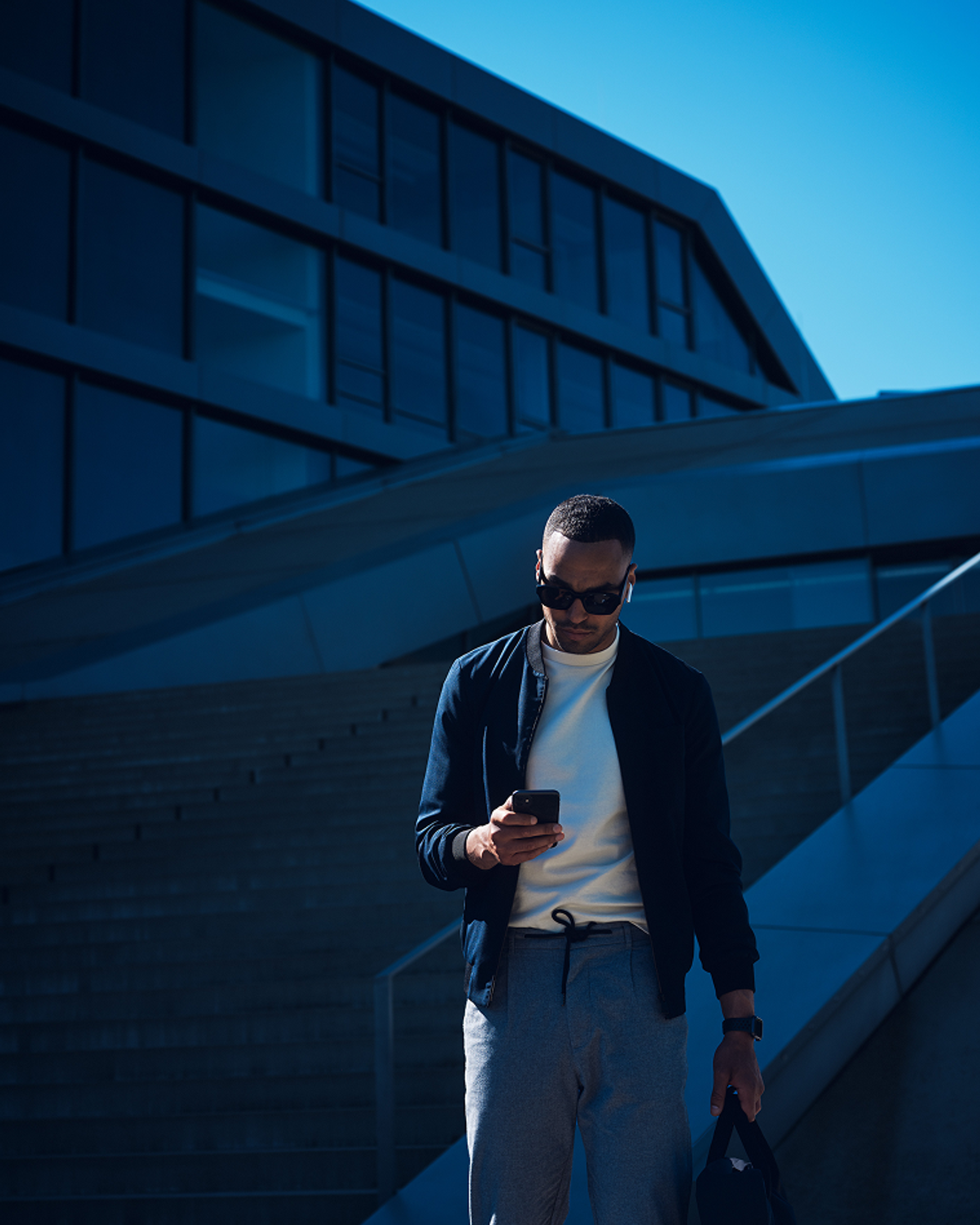 Mann mit Sonnenbrille und Tasche blickt auf Smartphone, blau getöntes Gegenlicht, urbane Architektur