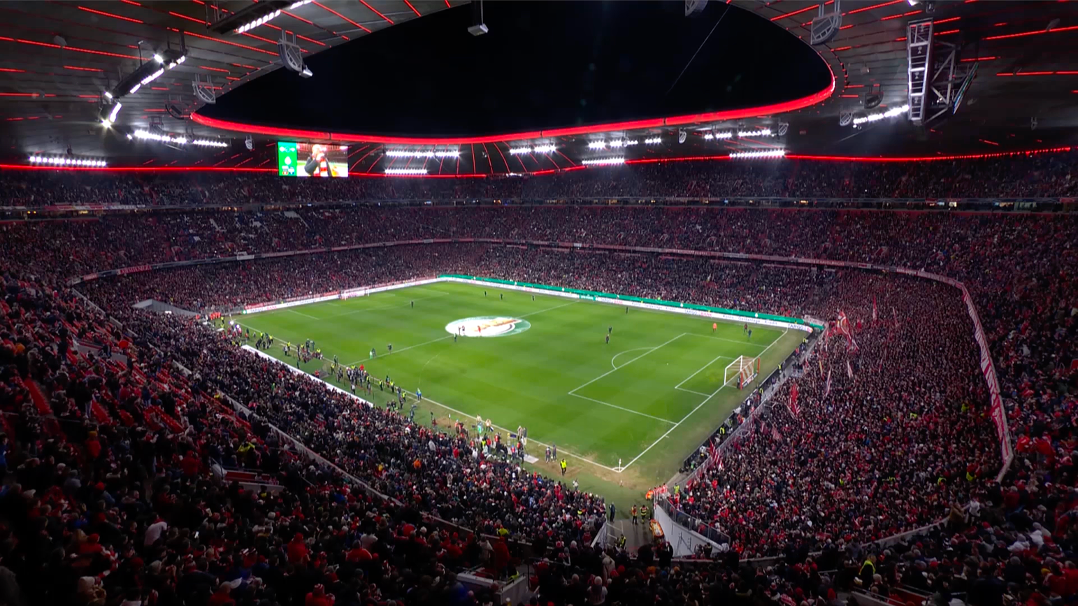 Gefülltes Fußballstadion bei Nacht mit Blick aufs Spielfeld und den beleuchteten Tribünen vor dem Spiel.