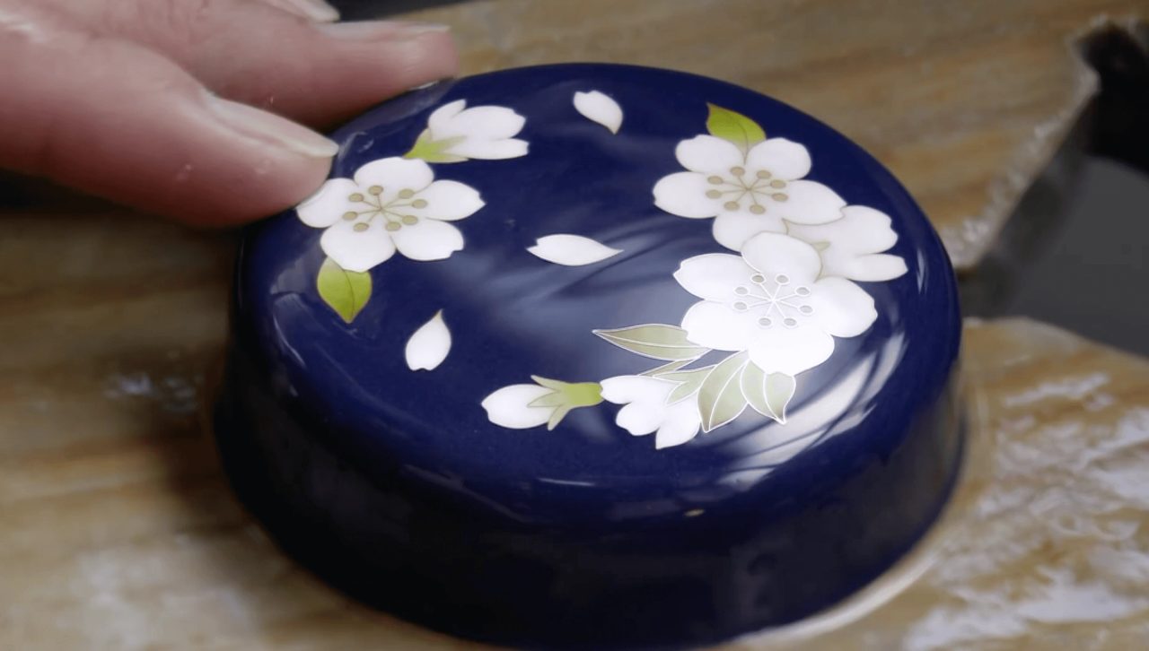 A hand steadying a deep blue enamelled dish decorated with white cherry blossoms and green leaves, resting face down on a wooden bench.