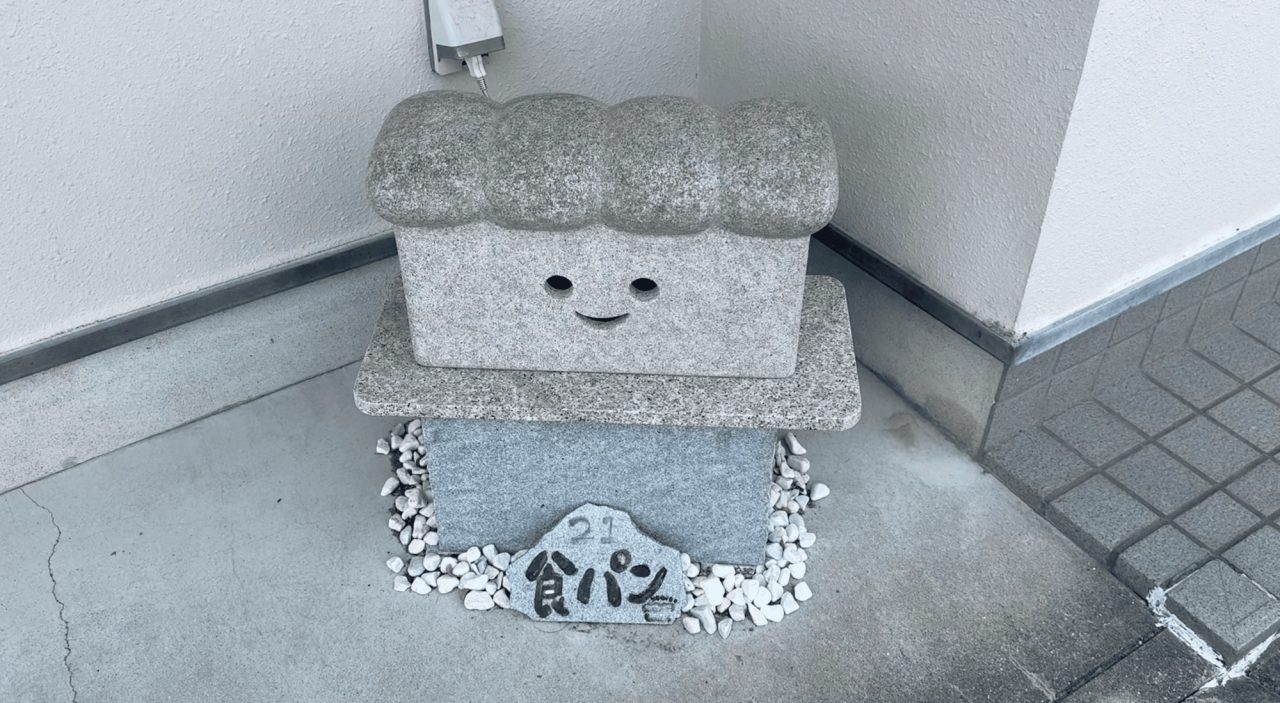 A granite ornament carved as a loaf of bread with a smiling face, set on a stone base outside a building, with white pebbles and a name plaque.