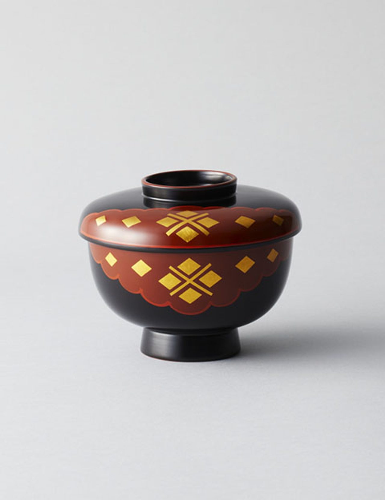 A small lidded lacquer bowl, black with a red band, decorated in gold diamonds and a four-diamond motif, on a pale grey background.