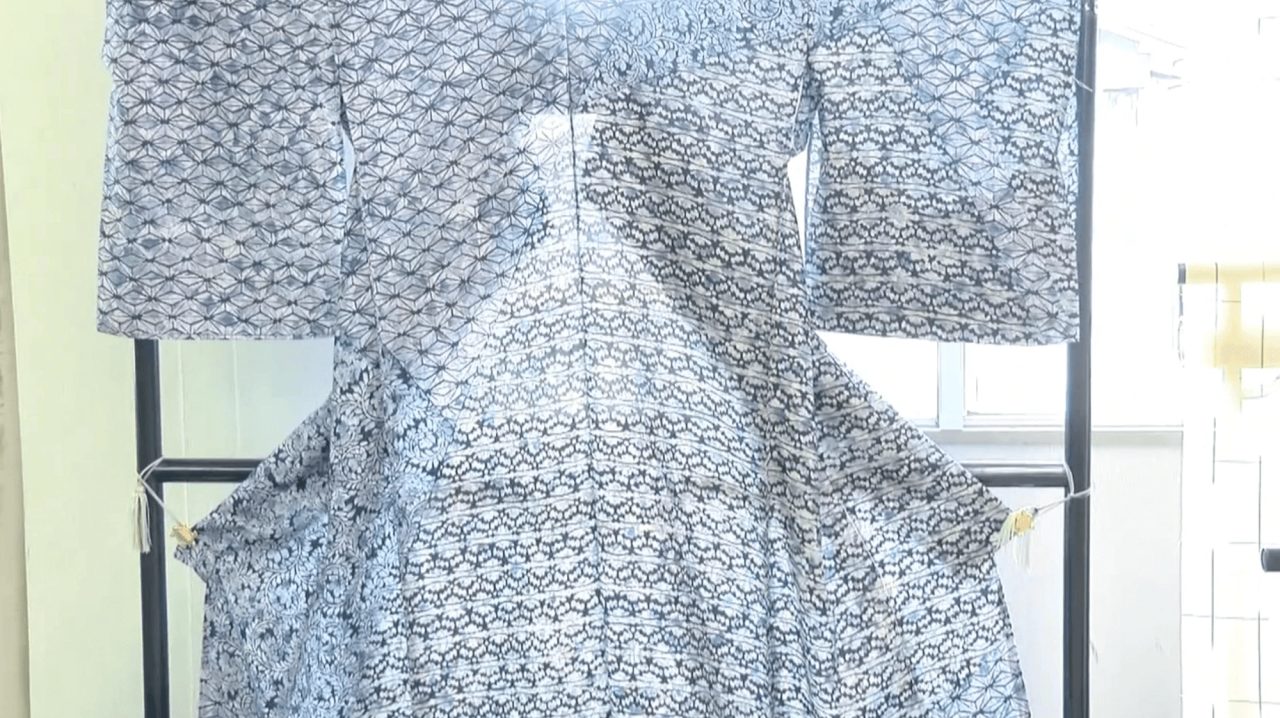 A kimono hung on a black rack in front of a window, woven in a dense indigo and white pattern across the whole garment.