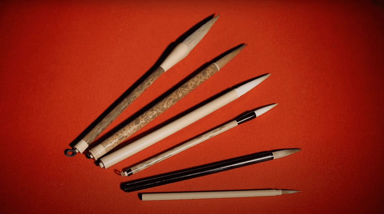 Six writing brushes fanned out on a deep red background, their handles in dark lacquer, pale bamboo and mottled wood.