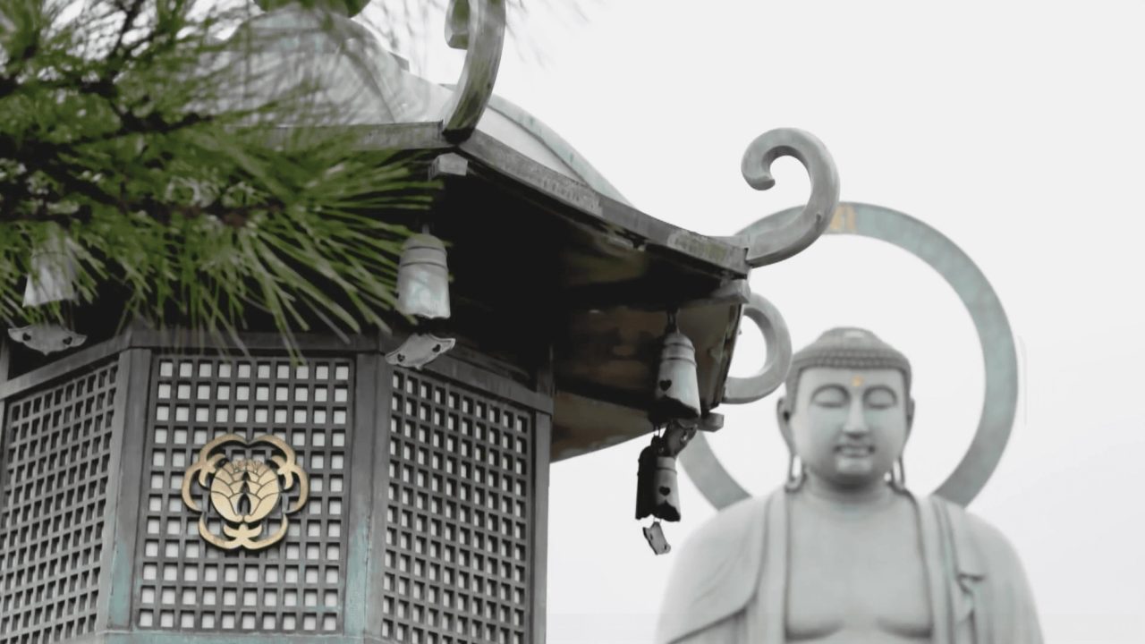 A bronze temple lantern with a curled finial and a chrysanthemum crest, a pine branch to the left and a large seated stone Buddha behind in mist.