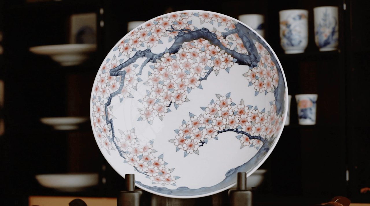 A wide porcelain dish painted with cherry blossom on grey branches in pink and white, displayed upright on a stand before a cabinet of cups.