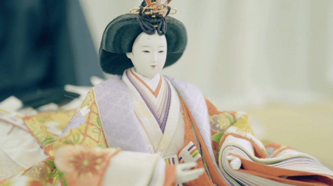 A seated doll with a white face and black lacquered hair in a court headdress, dressed in layered orange, gold and lavender silk.