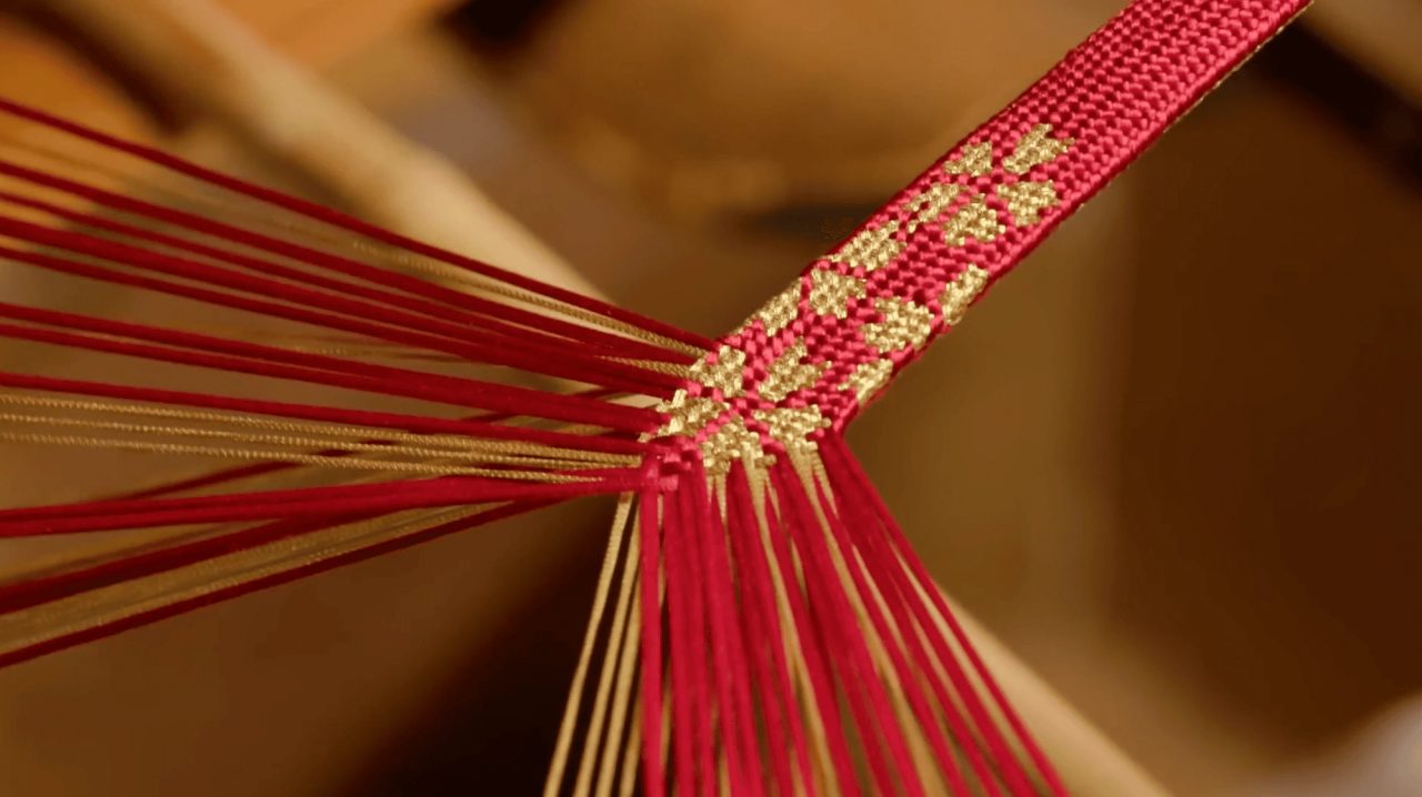 A braid of crimson and gold silk forming at the center of a braiding stand, with dozens of loose threads fanning out from the working point.