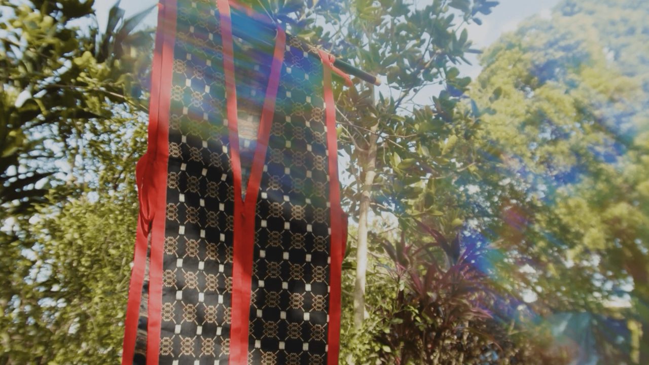 A length of dark cloth patterned with small pale geometric flowers, hanging outdoors with red ties, sunlight flaring through the trees behind.