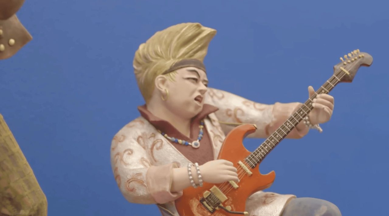 A painted clay figure of a young musician with blond swept-up hair, mid-performance on a red electric guitar, against a blue backdrop.