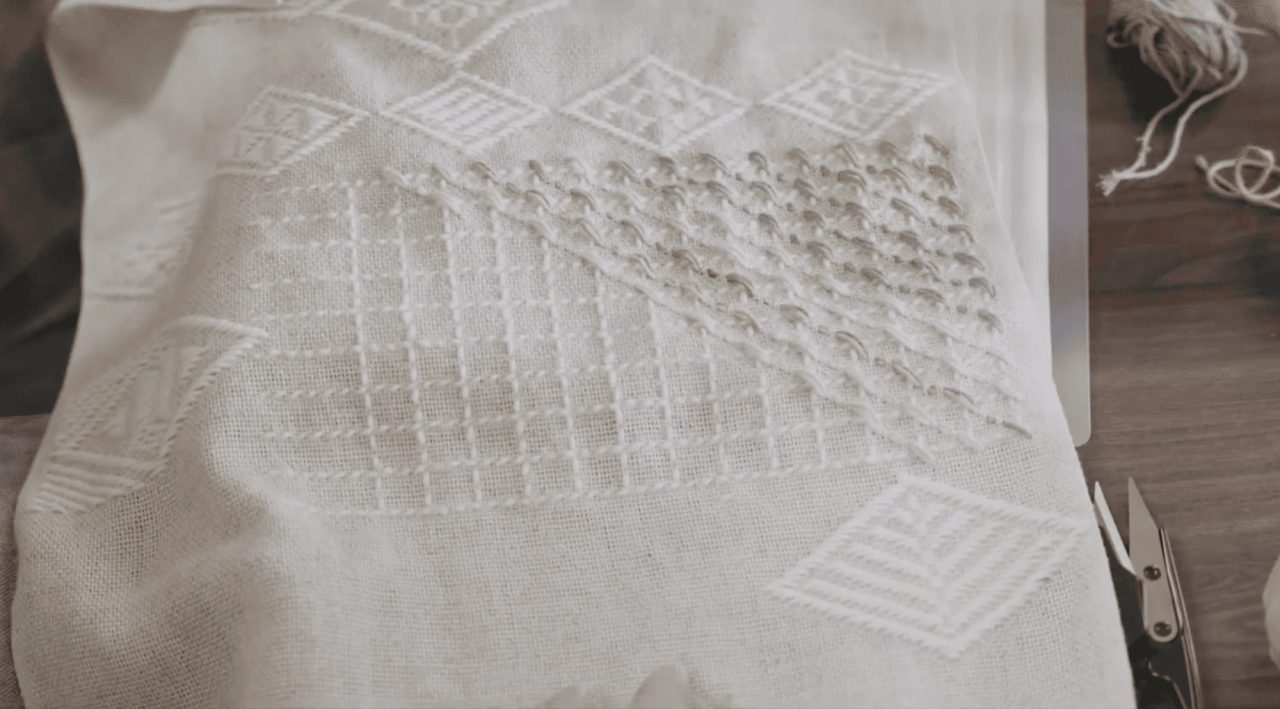Off-white linen stretched over a frame, embroidered in matching white thread with diamond and lattice patterns, scissors resting at the right.