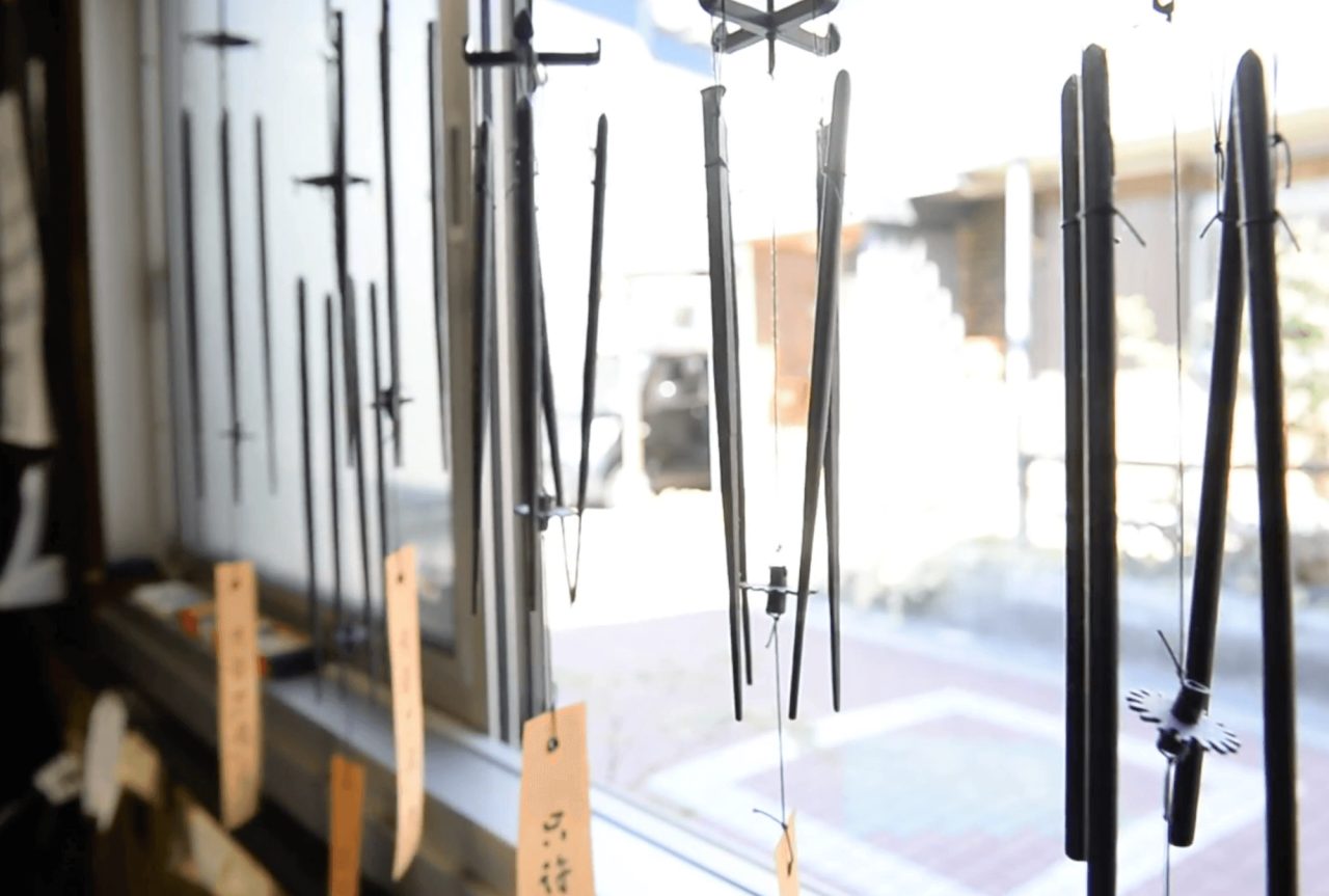 Slender forged iron rods hanging on threads in a shop window, some ending in small dark ornaments, with paper tags on the sill below.