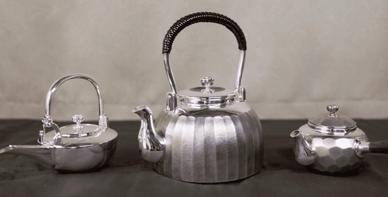 Three silver teapots on a dark cloth, one small and round, one large and fluted with a wrapped handle, one faceted with a side spout.
