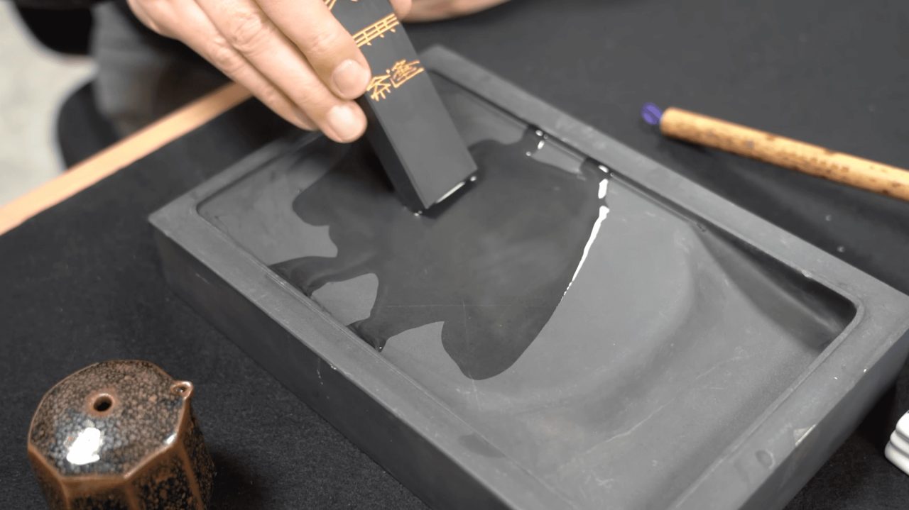 A hand rubbing a black inkstick with gold characters against a stone inkstone, grinding a pool of wet ink, a brush and water pot alongside.