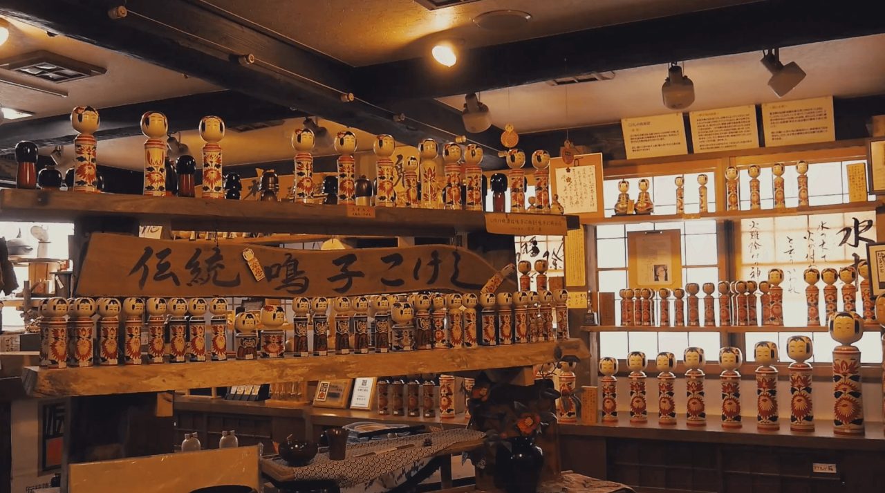 A shop interior with hundreds of turned wooden kokeshi dolls filling shelves and cabinets, a large painted wooden sign hanging above them.