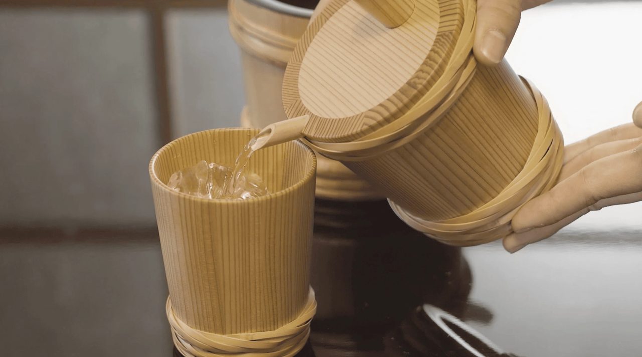 Hands tipping a cedar vessel bound with pale hoops to pour clear liquid into a matching cup on a dark surface.