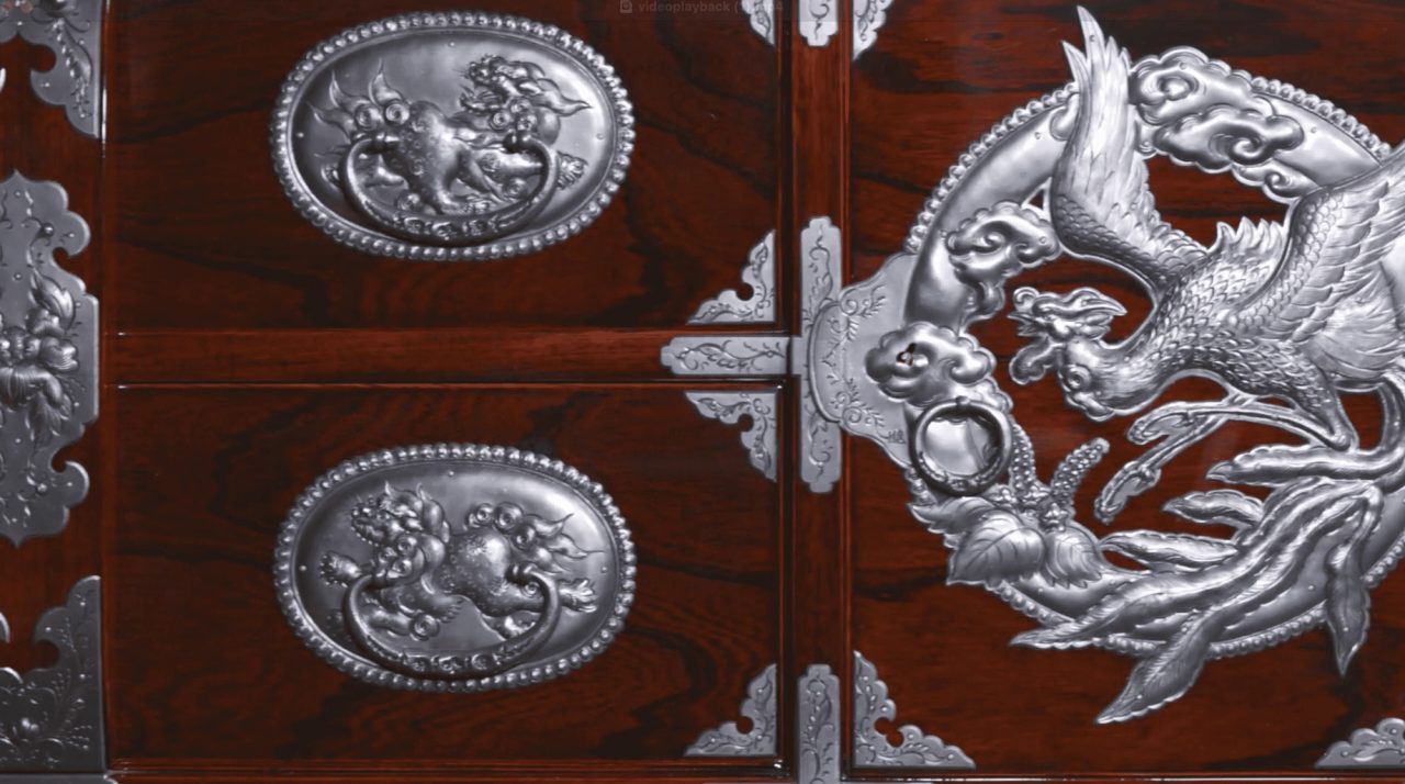 A close view of a dark lacquered chest, its drawers set with hammered metal plates showing lions in oval frames and a phoenix in flight.