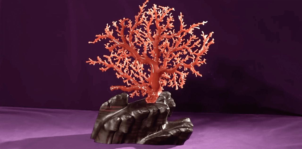 A branching piece of orange-red coral mounted upright on a dark carved stone base, against a purple backdrop.