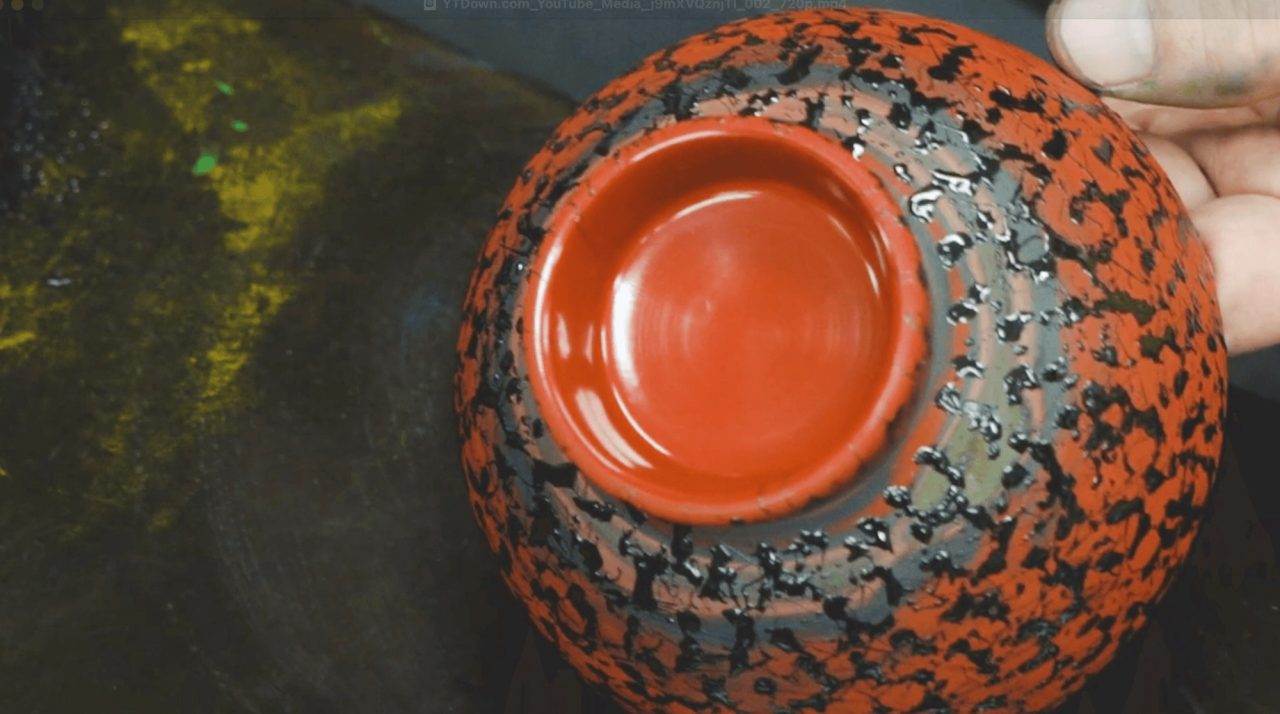 A bowl held in one hand and seen from above, its lacquer surface a mottled pattern of orange, black and grey around a plain orange well.