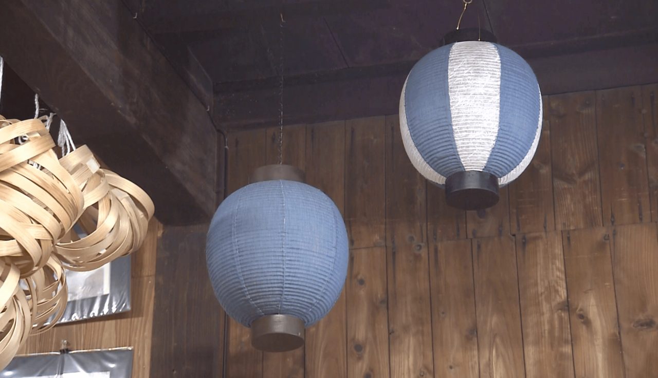 Two round paper lanterns hanging from a dark beam in a wooden building, one plain blue, one blue with a wide white panel.
