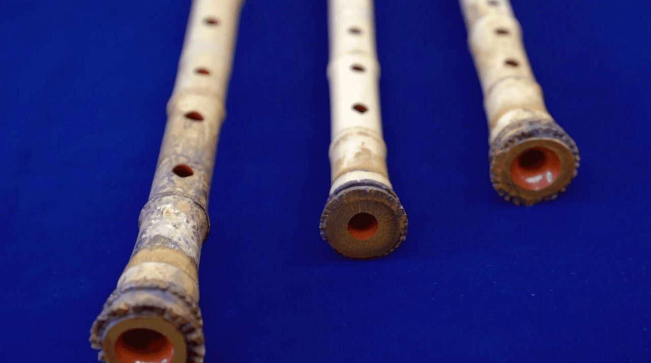 Three bamboo flutes laid on blue cloth, their finger holes in a line and their root ends flaring around a lacquered red bore.