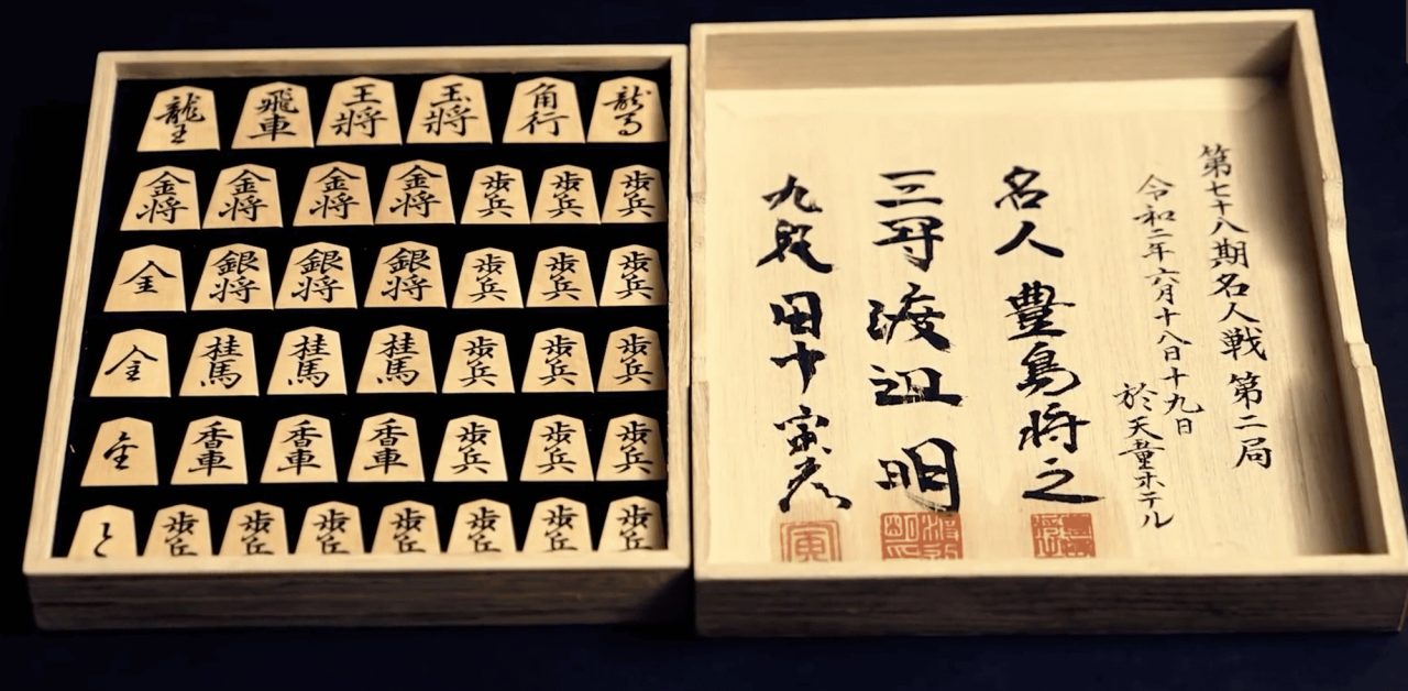 A wooden box of shogi pieces arranged in rows with their characters brushed in black, beside a lid inscribed in calligraphy with red seals.