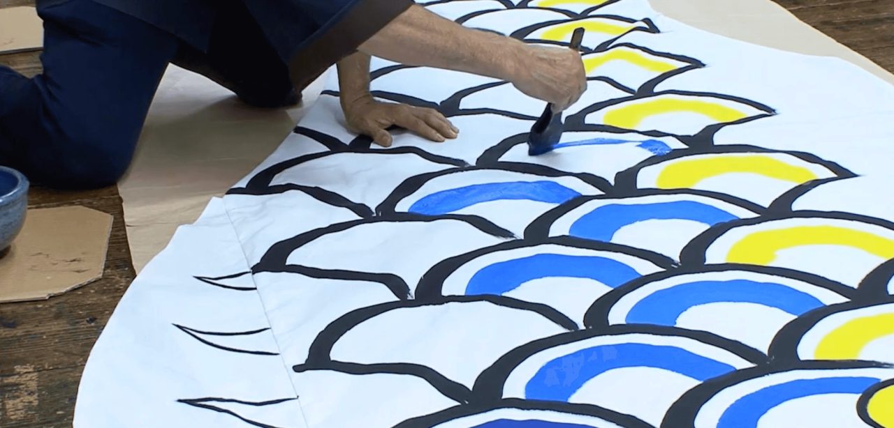 A person kneeling on a workshop floor, brushing blue paint into the outlined scale pattern of a large white carp streamer.