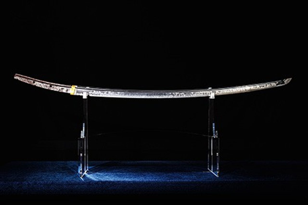 A long curved sword blade mounted horizontally on two clear stands, lit against a black background so the temper line shows along its edge.