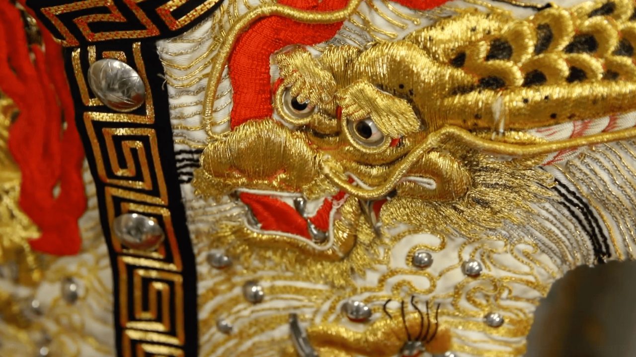 A close view of a gold-embroidered dragon head on a costume, worked in raised metallic thread over red and white silk with silver studs.