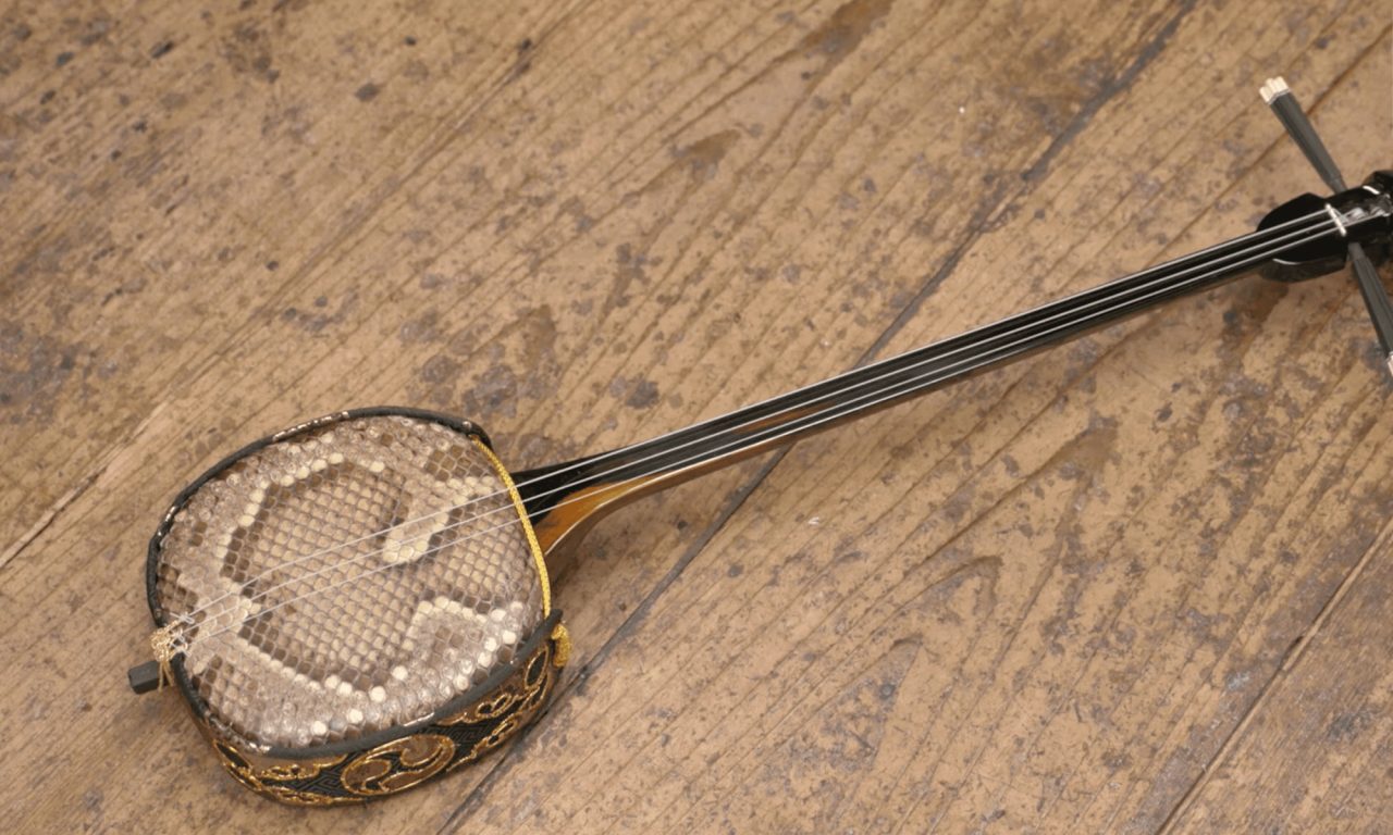 A three-stringed sanshin lying on a weathered wooden floor, its round body covered in snakeskin, with a long black neck.