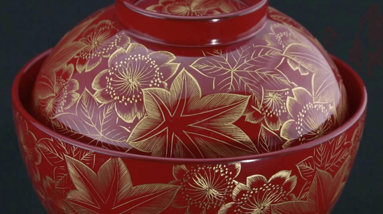 A lidded red lacquer bowl decorated in gold with maple leaves and camellia flowers across the lid and body, on a dark background.