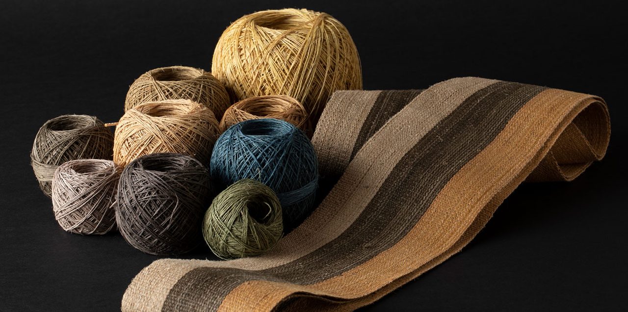 Ten balls of hand-spun bark thread in ochre, teal, olive and grey, beside a folded band of striped woven cloth on a black surface.