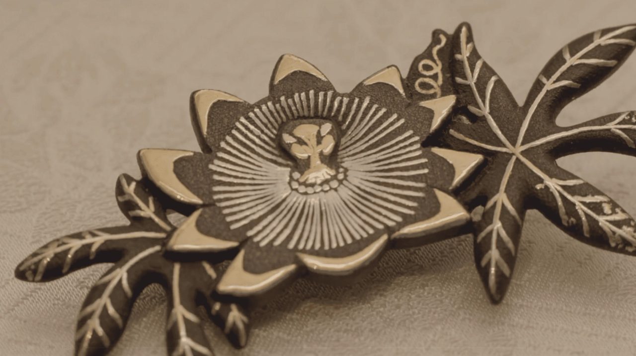 A dark metal ornament of a flower and leaves, its surface inlaid with fine silver lines radiating from the center, on pale cloth.